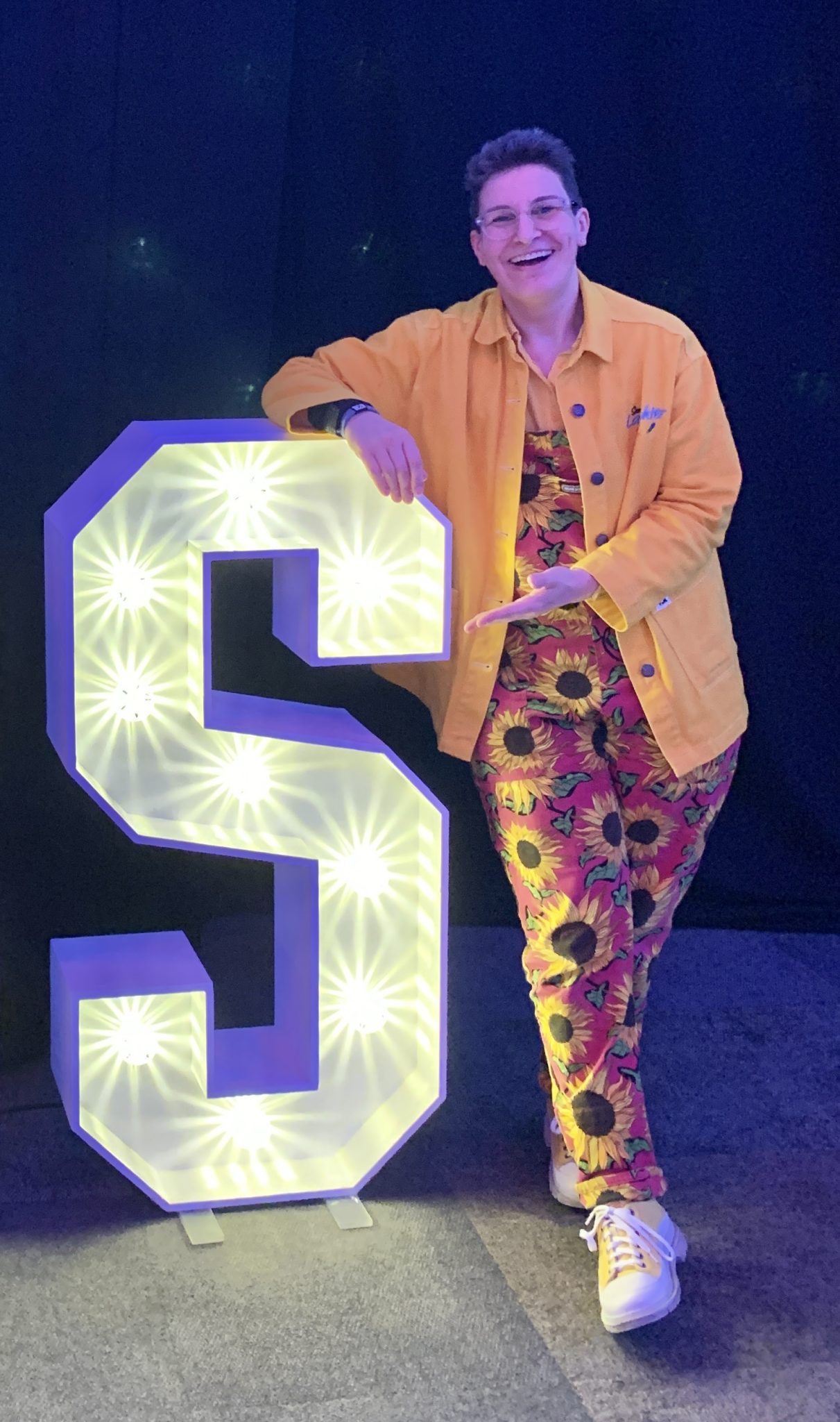 Sara Kay woman with sunflower dungarees and yellow jacket next to an illuminated letter S